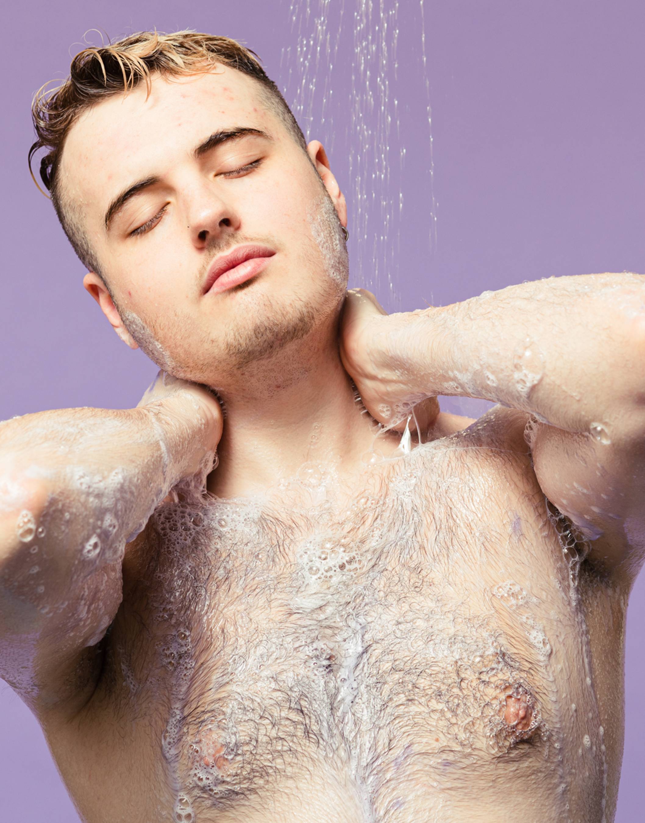 A person with eyes closed relaxes under a stream of shower water, massaging their neck with their hands. Fluffy sudsy lather covers their hairy chest and arms, set against a solid pastel purple background.