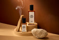 The image shows a warm, bark-brown background. In the foreground are the two different-sized bottles of the Vegen Leather Jacket perfume on a small, stacked pedestal with a smoking piece of wood and a porous rock beside them.